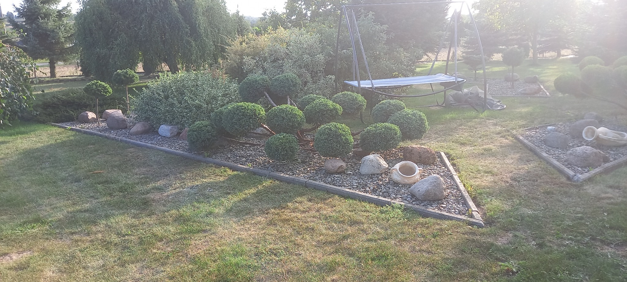 Ogród z formowanymi krzewami w kształcie kul, otoczony kamieniami i obrzeżem, uzupełniony dekoracyjnymi amforami i huśtawką w tle.