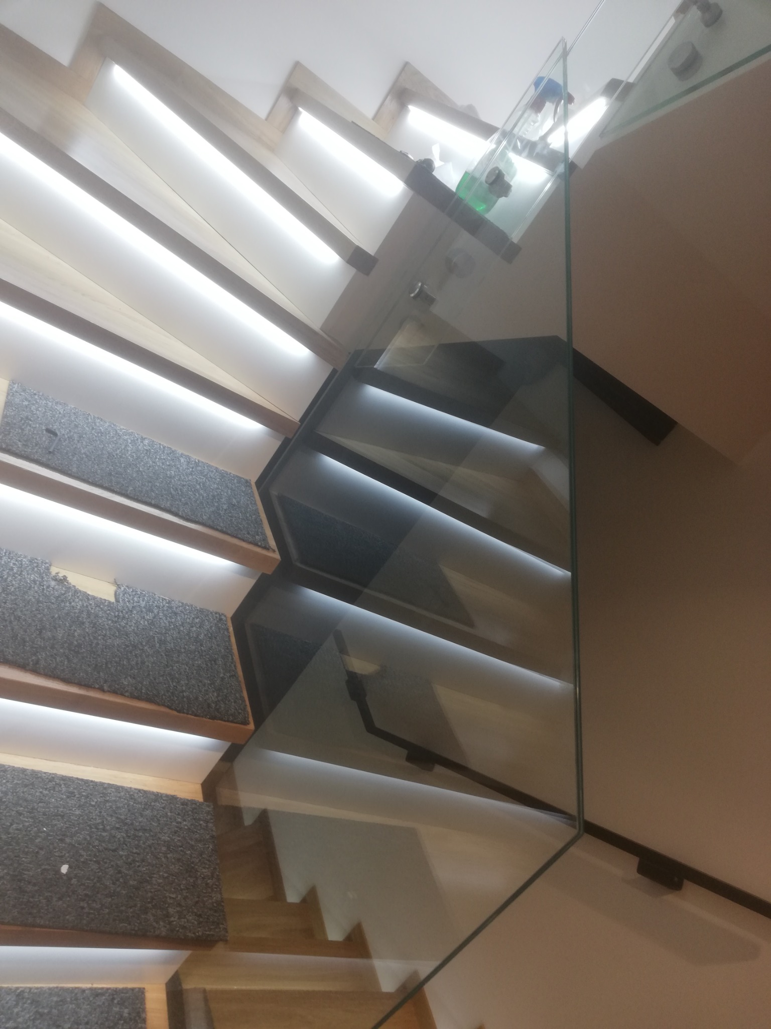 Nowoczesne schody z drewnianymi stopniami pokrytymi szarym dywanem i szklaną balustradą, widok z dołu, oświetlenie LED pod stopniami.