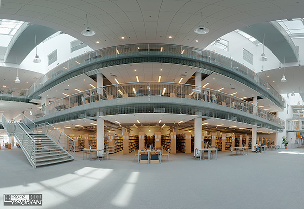 Panoramiczny widok wnętrza nowoczesnej biblioteki z trzema poziomami, metalowymi balustradami, przeszklonym dachem i rzędami regałów z książkami.