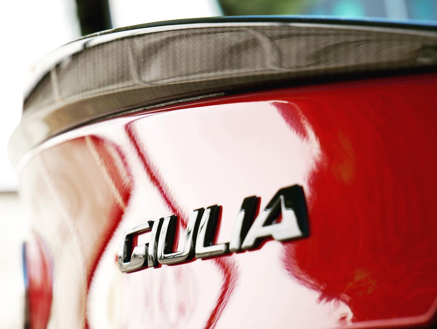 Czerwony lakierowany bagażnik samochodu Alfa Romeo Giulia z chromowanym napisem modelu i karbonowym spojlerem, widoczne refleksy świetlne.