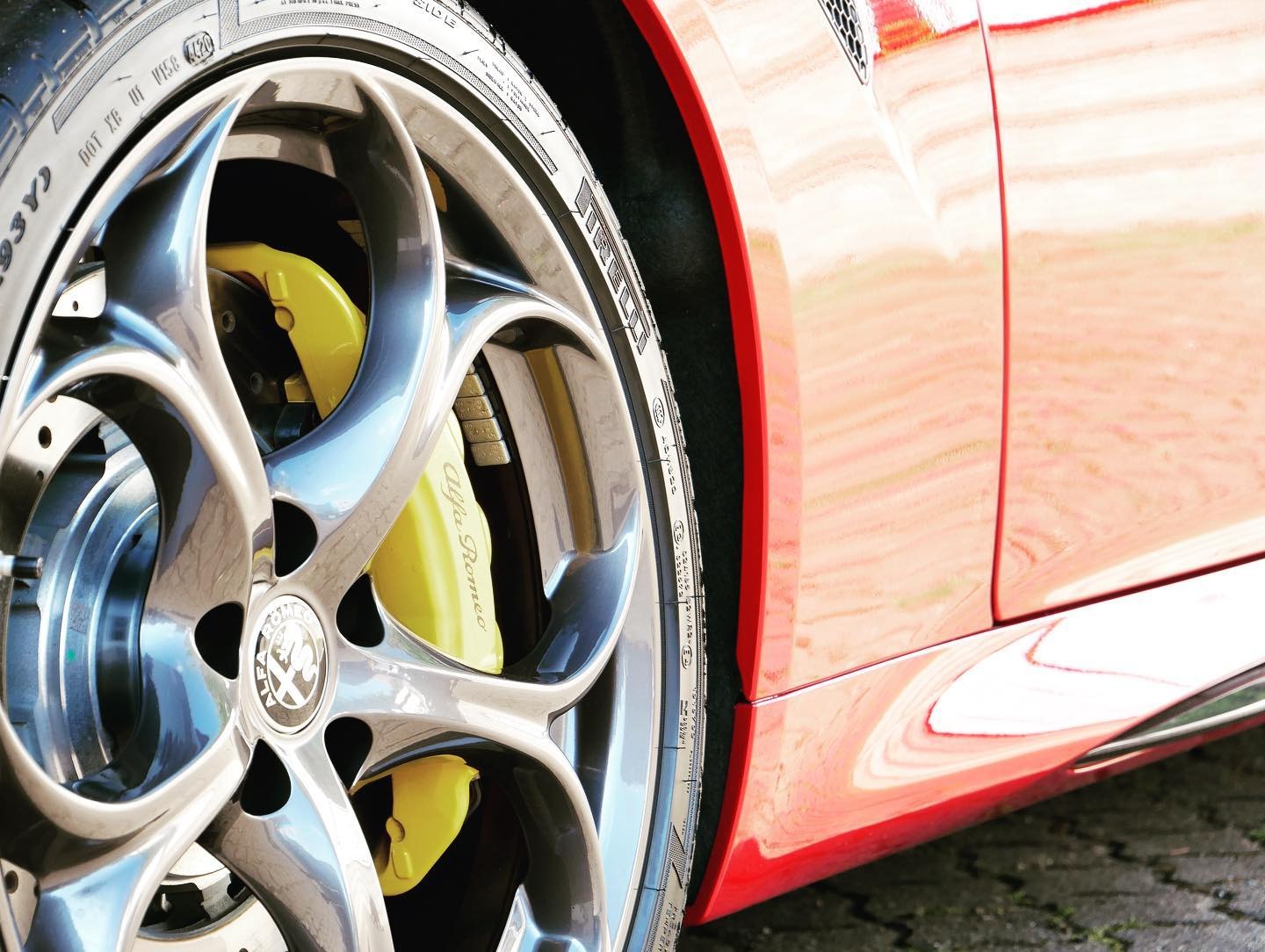 Błyszczące, wieloramienne felgi aluminiowe z żółtymi zaciskami hamulcowymi Alfa Romeo na tle karoserii w odcieniach czerwieni.