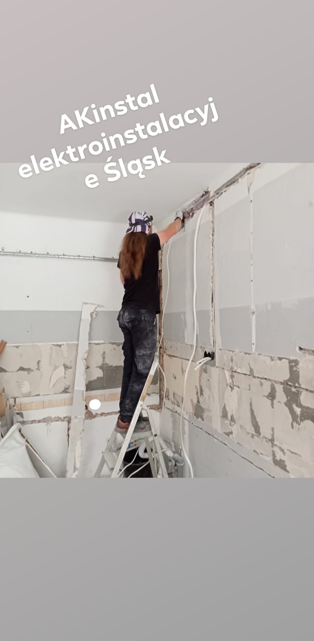 Elektryk na drabinie montuje kable w puszce elektrycznej w remontowanym pomieszczeniu z częściowo położonymi płytami kartonowo-gipsowymi, widoczne przewody i metalowe korytko kablowe pod sufitem.