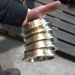 Ręka trzymająca stos obrobionych, metalowych pierścieni z kołnierzami, widoczne ślady obróbki, tło warsztatowe.