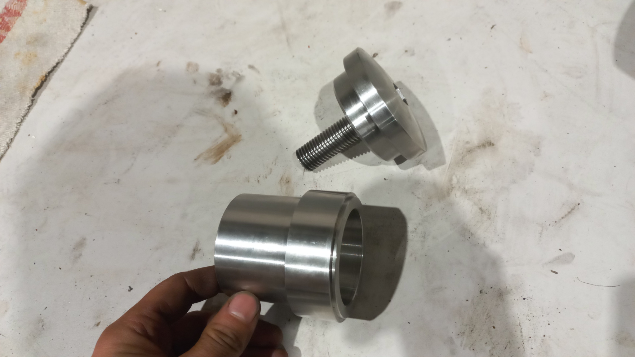 Dwa precyzyjnie wykonane, metalowe elementy cylindryczne z gwintem, leżące na zabrudzonym blacie roboczym, trzymane w dłoni.