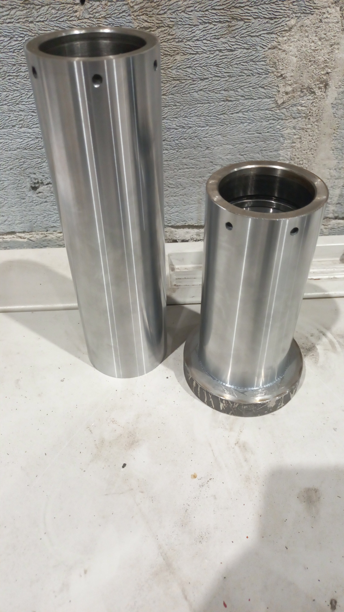 Dwa cylindryczne, metalowe elementy z otworami, jeden z podstawą, widoczne ślady obróbki.