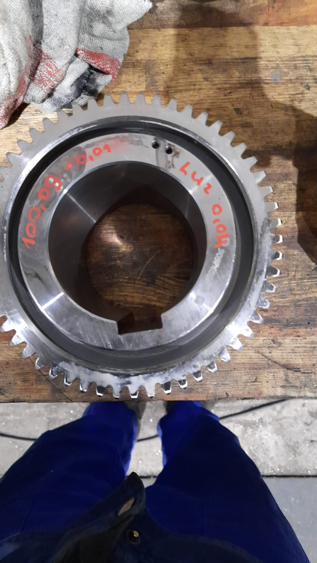 Metalowa zębatka z naniesionymi czerwonymi oznaczeniami leży na drewnianym blacie warsztatowym, obok widoczna zabrudzona szmatka i fragment niebieskiego kombinezonu.