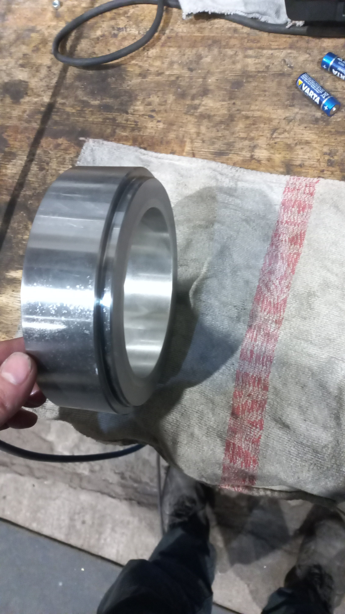 Metalowy pierścień z rowkiem, trzymany w dłoni, leżący na szarej szmatce z czerwonym paskiem, w tle roboczy stół z narzędziami i bateriami.