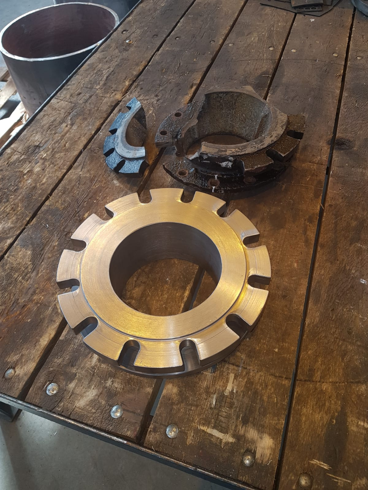 Metalowy pierścień z wycięciami, fragmenty zużytych elementów maszyny i rura na tle drewnianego stołu warsztatowego.