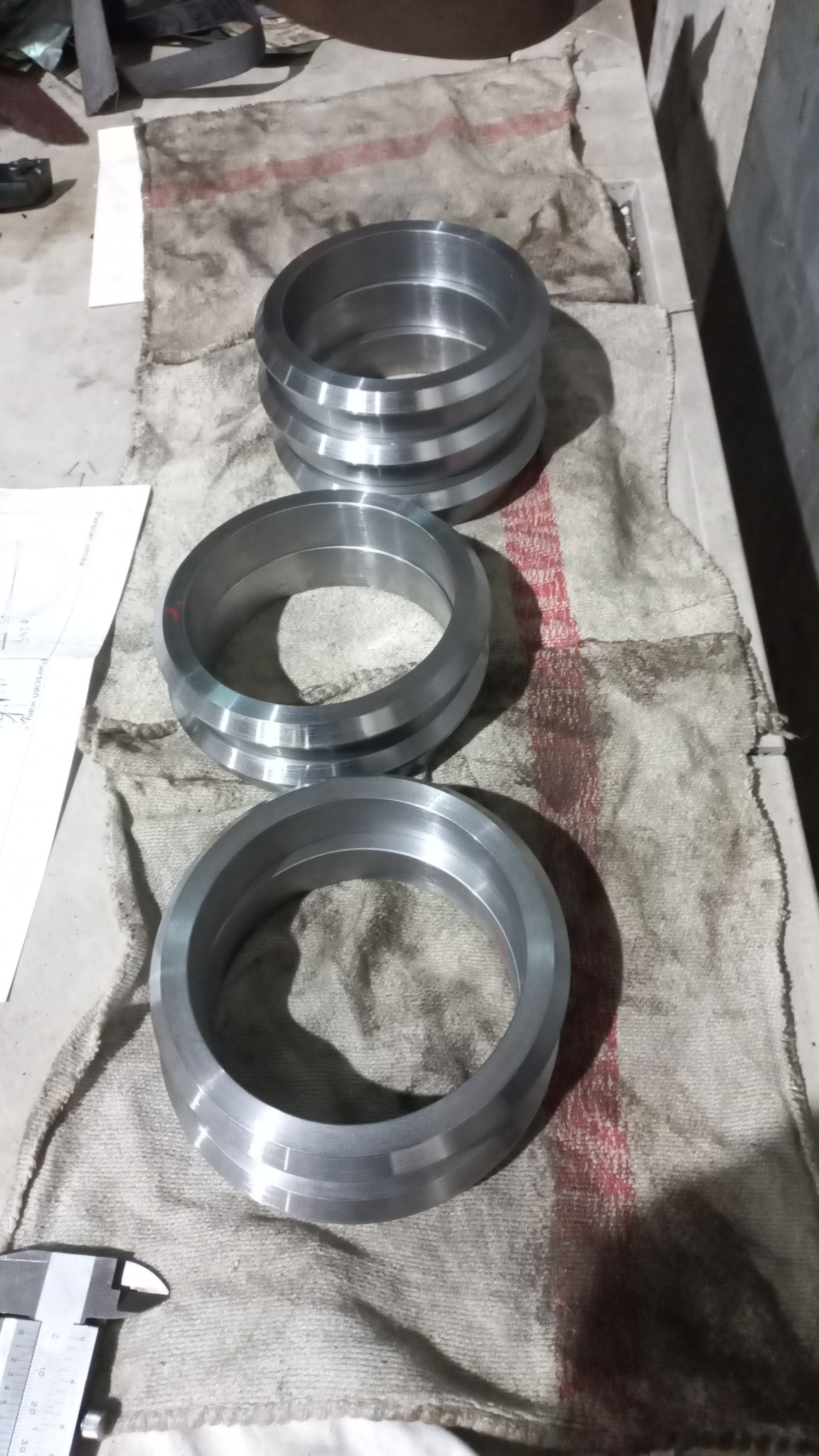 Trzy metalowe pierścienie o różnej średnicy, ułożone jeden na drugim, leżą na brudnej szmacie, obok leży suwmiarka i częściowo widoczny jest rysunek techniczny.