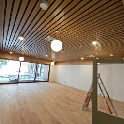 ELEKTRO - INSTAL ŁUKASZ KOLADA - Wnętrze biura w trakcie remontu: drewniana podłoga, sufit z listew, wiszące okrągłe lampy, aluminiowa drabina oparta o ścianę, duże okno z widokiem na plac budowy na zewnątrz.