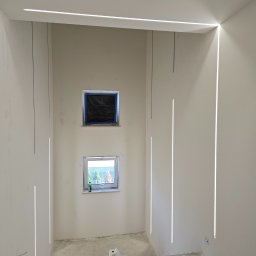ELEKTRO - INSTAL ŁUKASZ KOLADA - Wnętrze w trakcie remontu z zamontowanym nowoczesnym oświetleniem liniowym LED w ścianach i na suficie. Dwa okna, jedno zakryte folią malarską, surowa podłoga betonowa, widoczne ślady prac...