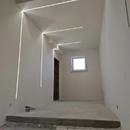 Wnętrze w trakcie remontu z zamontowanym liniowym oświetleniem LED w ścianach i suficie, surowy beton na schodach i podłodze, widoczne puszki elektryczne i okno z widokiem na zewnątrz.