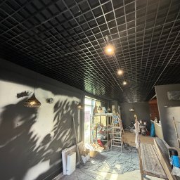 ELEKTRO - INSTAL ŁUKASZ KOLADA - Wnętrze w trakcie remontu: czarny sufit kasetonowy z oświetleniem punktowym, ściany pomalowane na szaro. Widoczne rusztowania i narzędzia malarskie.