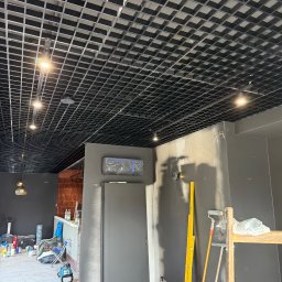 ELEKTRO - INSTAL ŁUKASZ KOLADA - Sufit rastrowy z oświetleniem punktowym w lokalu komercyjnym w trakcie remontu. Widoczne drabina, poziomica i narzędzia malarskie.