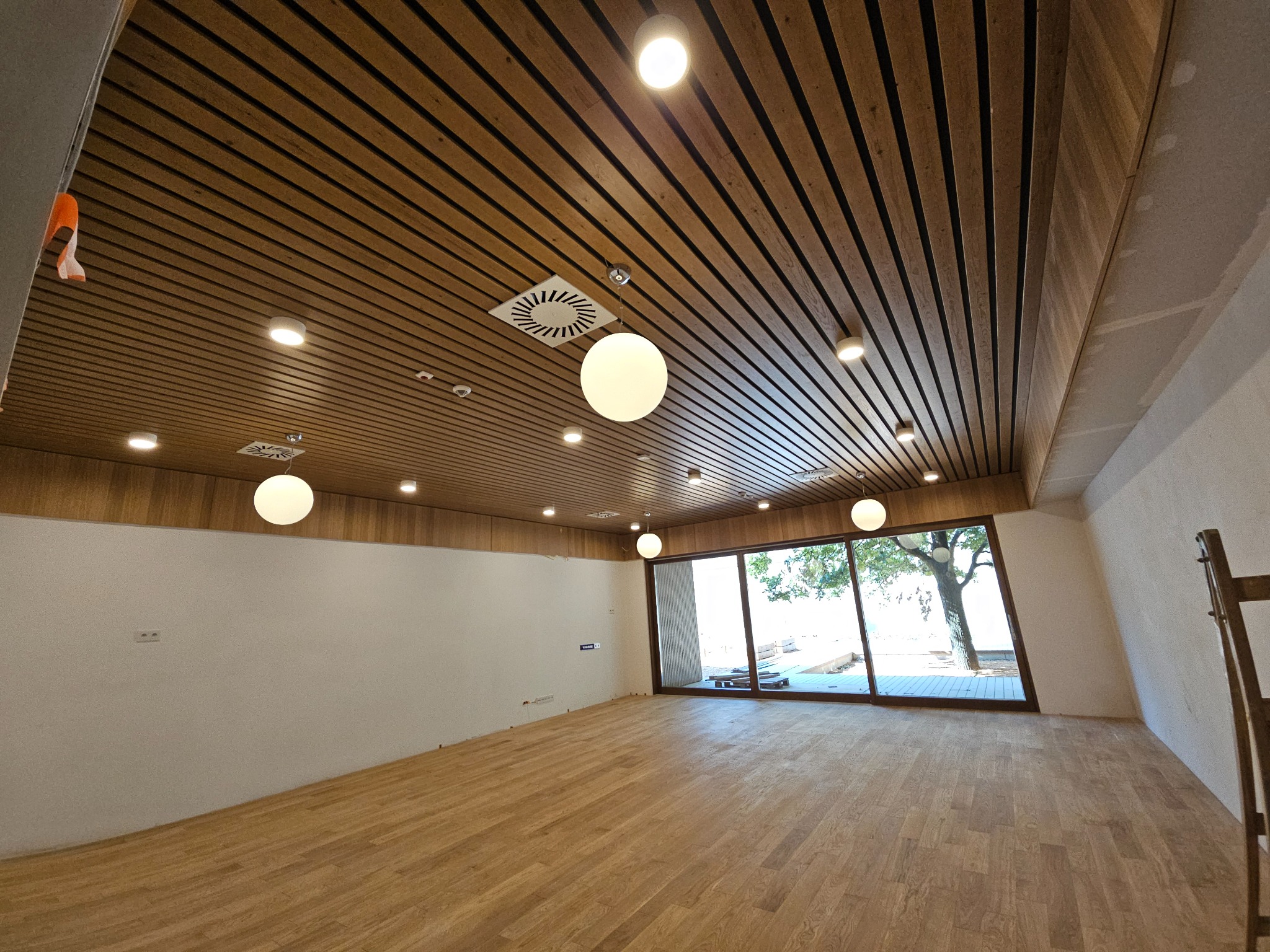 Wnętrze biura po remoncie z drewnianym parkietem i sufitem z listew, widoczne okna z widokiem na drzewo, zamontowane oświetlenie punktowe i wiszące lampy kuliste.