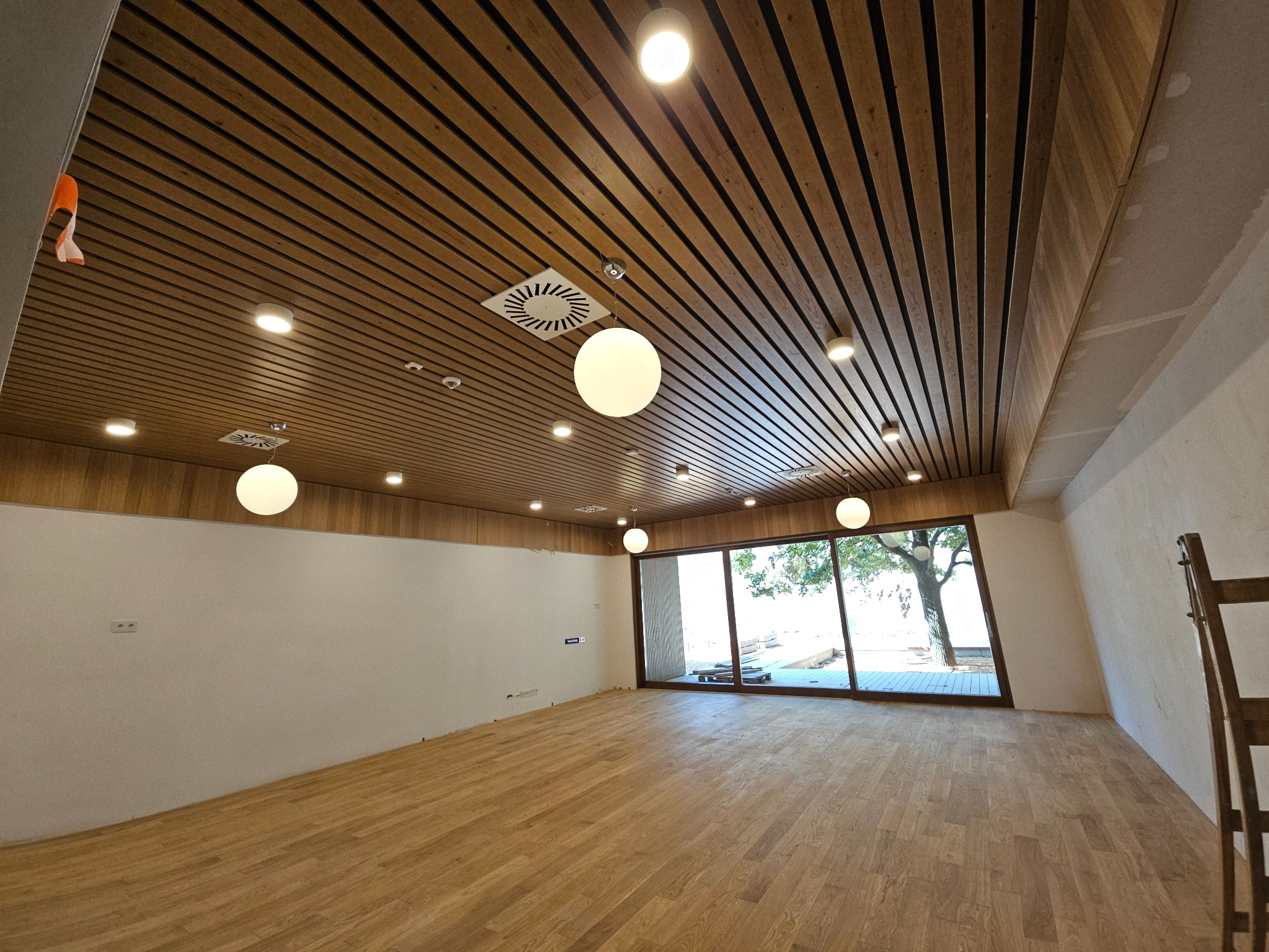 Przestronne wnętrze biurowe z drewnianym parkietem i podwieszanym sufitem z oświetleniem punktowym oraz kulistymi lampami wiszącymi, duże okno z widokiem na zewnątrz, ściany w trakcie wykończenia.