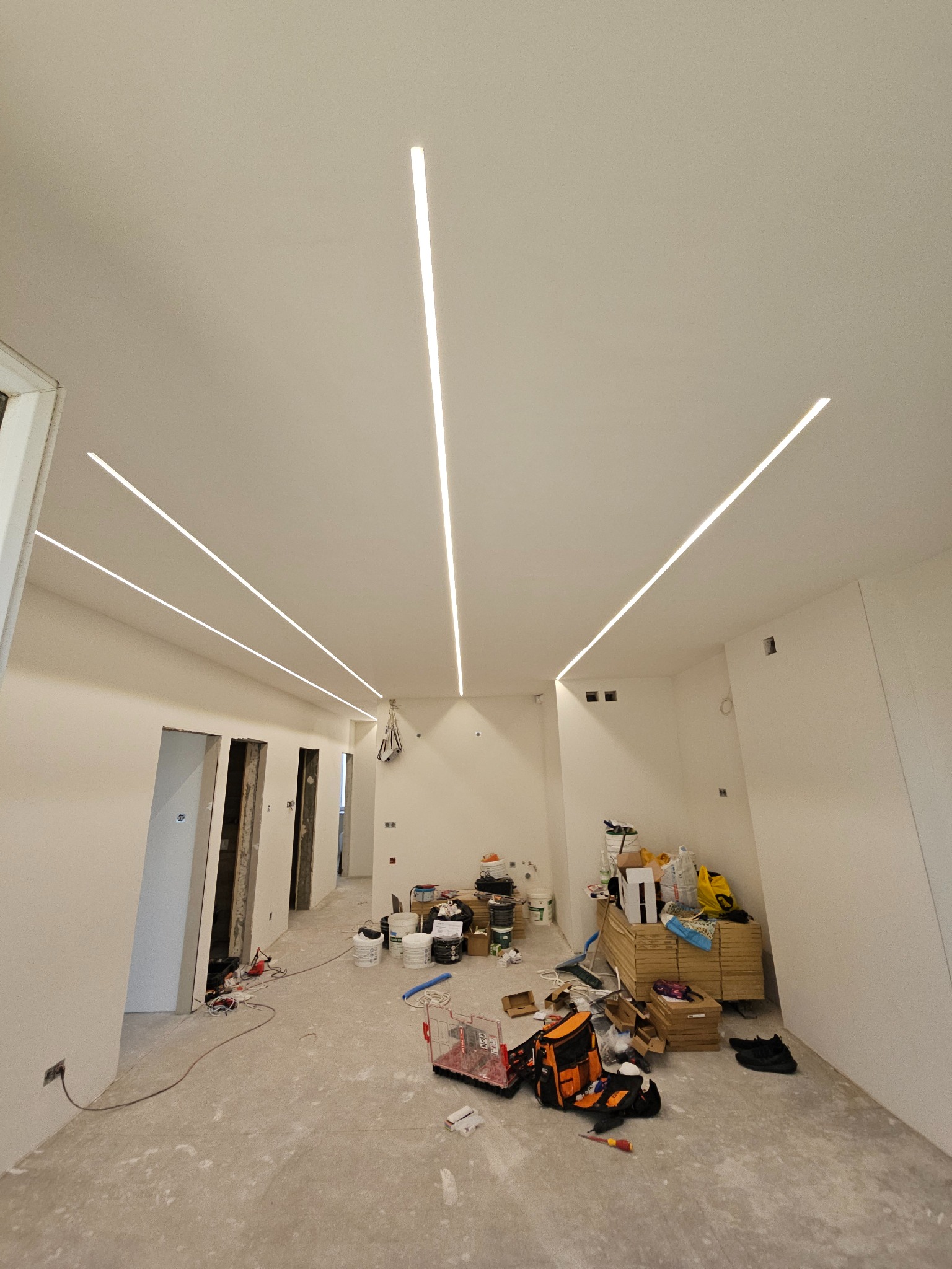 Wnętrze w trakcie remontu z zamontowanym liniowym oświetleniem LED w suficie, widoczne narzędzia i materiały budowlane na podłodze.