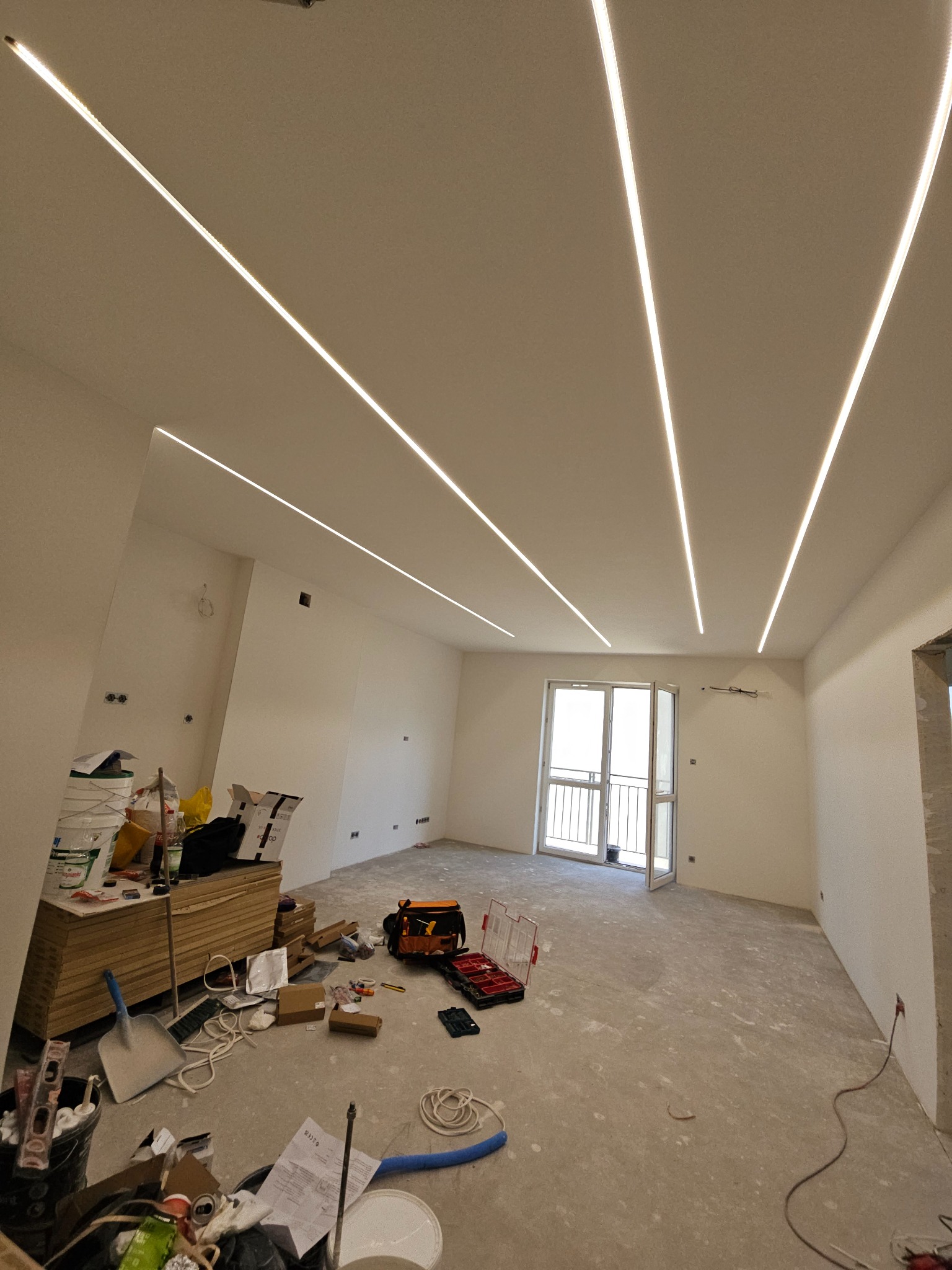 Wnętrze w trakcie remontu z zamontowanym liniowym oświetleniem LED w suficie, widoczne narzędzia i materiały budowlane, surowe ściany i podłoga, otwarte drzwi balkonowe.