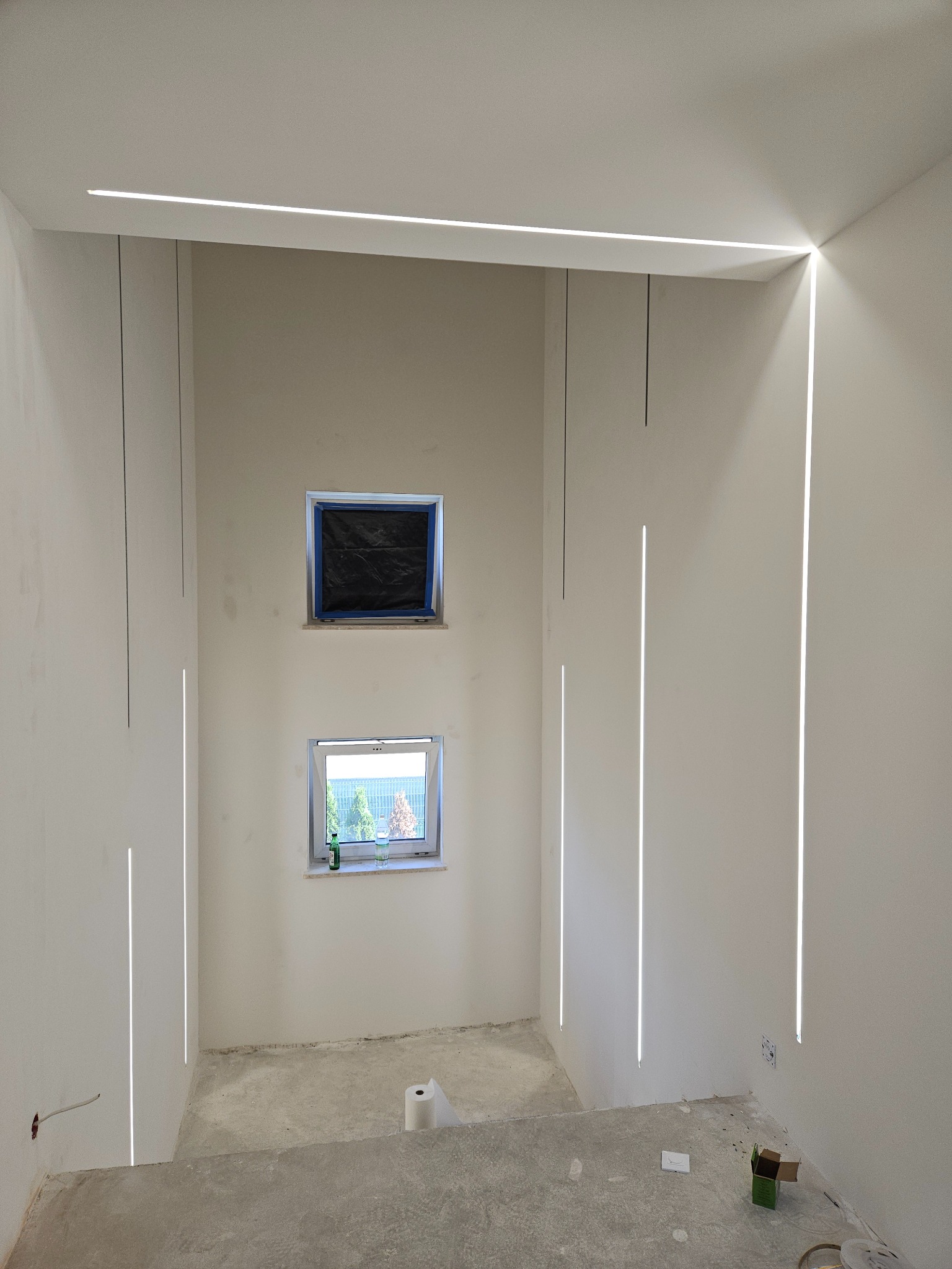 Wnętrze w trakcie remontu z zamontowanym nowoczesnym oświetleniem liniowym LED w ścianach i na suficie. Dwa okna, jedno zakryte folią malarską, surowa podłoga betonowa, widoczne ślady prac...