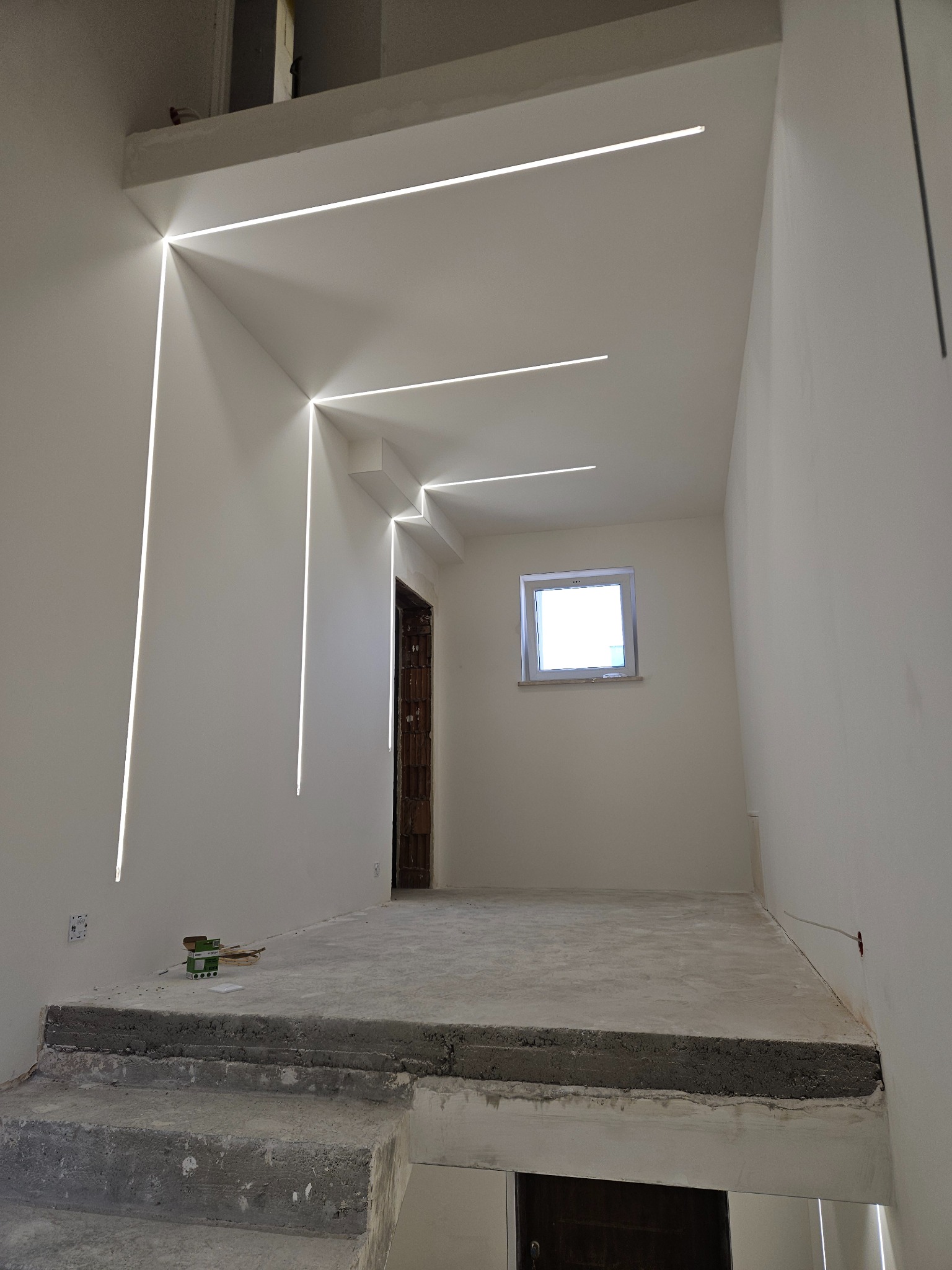 Wnętrze w trakcie remontu z zamontowanym liniowym oświetleniem LED w ścianach i suficie, surowy beton na schodach i podłodze, widoczne puszki elektryczne i okno z widokiem na zewnątrz.