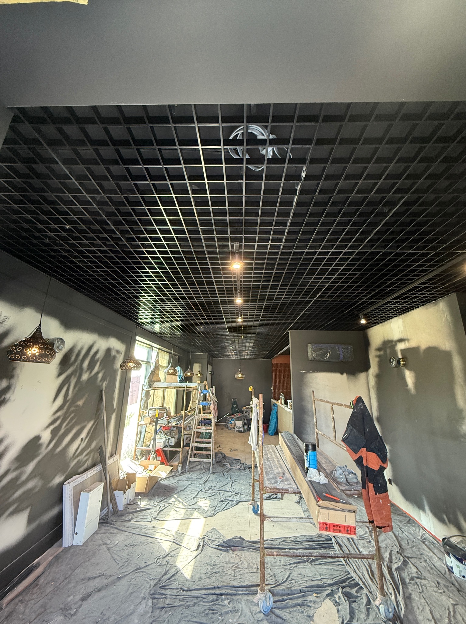Sufit rastrowy w trakcie montażu oświetlenia w szarym wnętrzu. Widoczne rusztowania i narzędzia, co sugeruje etap prac wykończeniowych. Lampy dekoracyjne w tle.