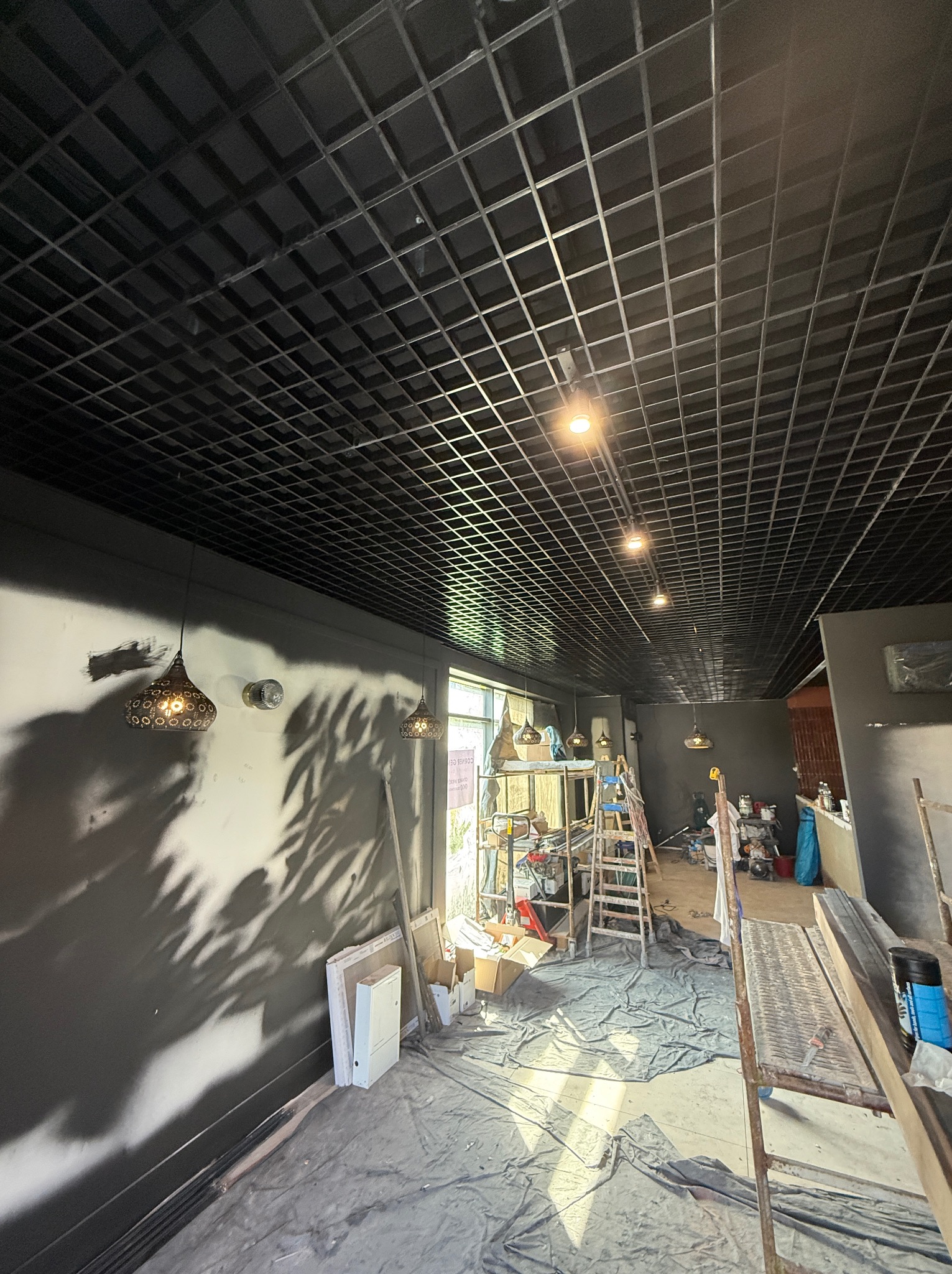 Wnętrze w trakcie remontu: czarny sufit kasetonowy z oświetleniem punktowym, ściany pomalowane na szaro. Widoczne rusztowania i narzędzia malarskie.