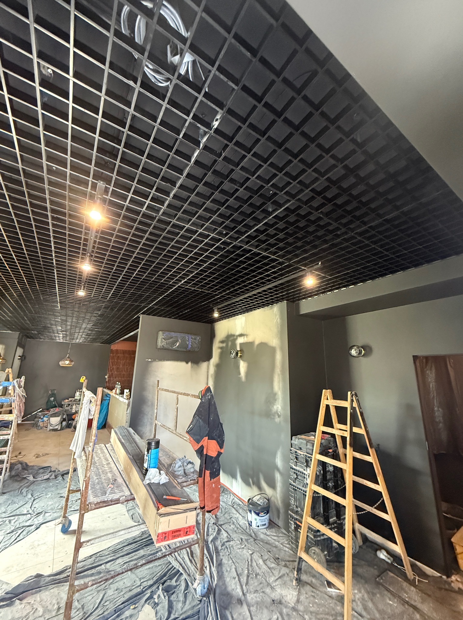 Sufit rastrowy z zamontowanym oświetleniem w trakcie remontu biura, widoczne drabiny i zabezpieczenia podłogi, szare ściany i lampy ścienne.