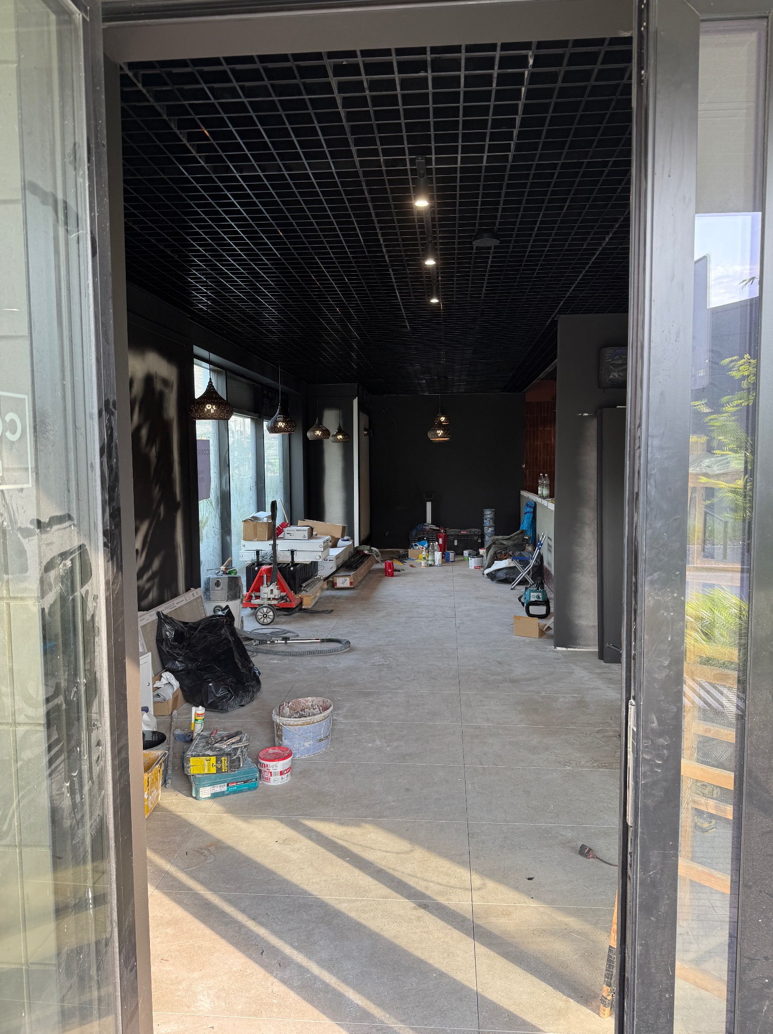 Wnętrze biura w trakcie remontu: czarne ściany, podwieszany sufit z oświetleniem, widoczne narzędzia i materiały budowlane, otwarte drzwi wejściowe.