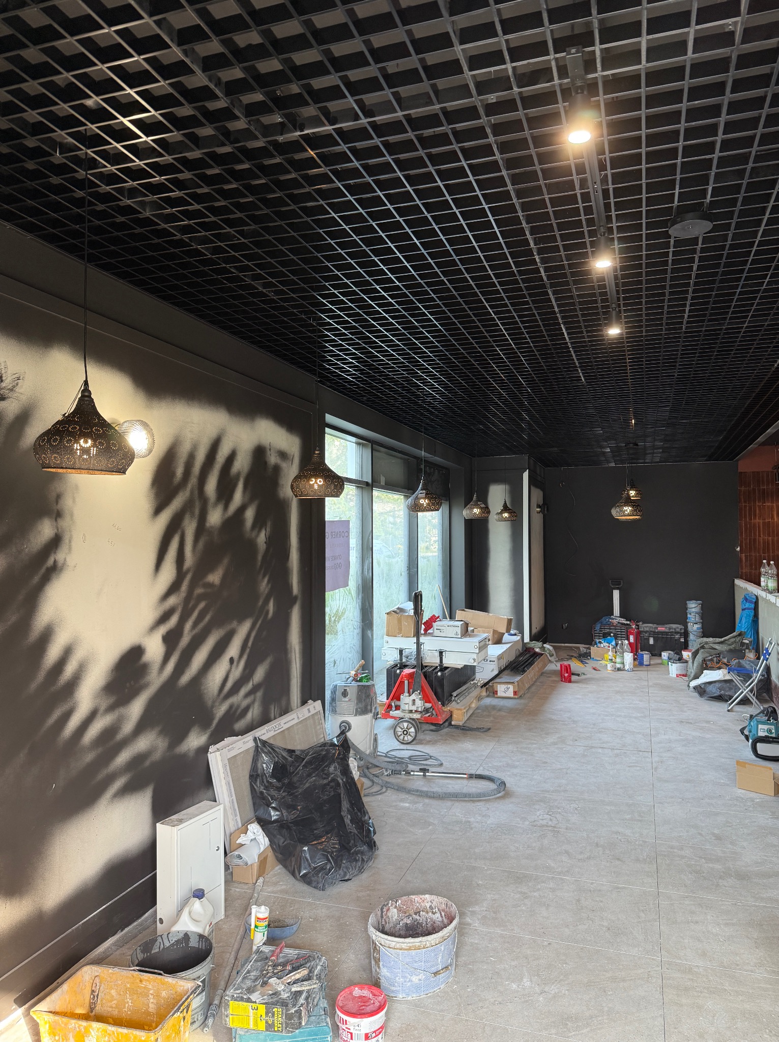 Wnętrze biura w trakcie remontu z zamontowanym oświetleniem sufitowym i wiszącymi lampami. Na podłodze widoczne materiały budowlane i narzędzia.