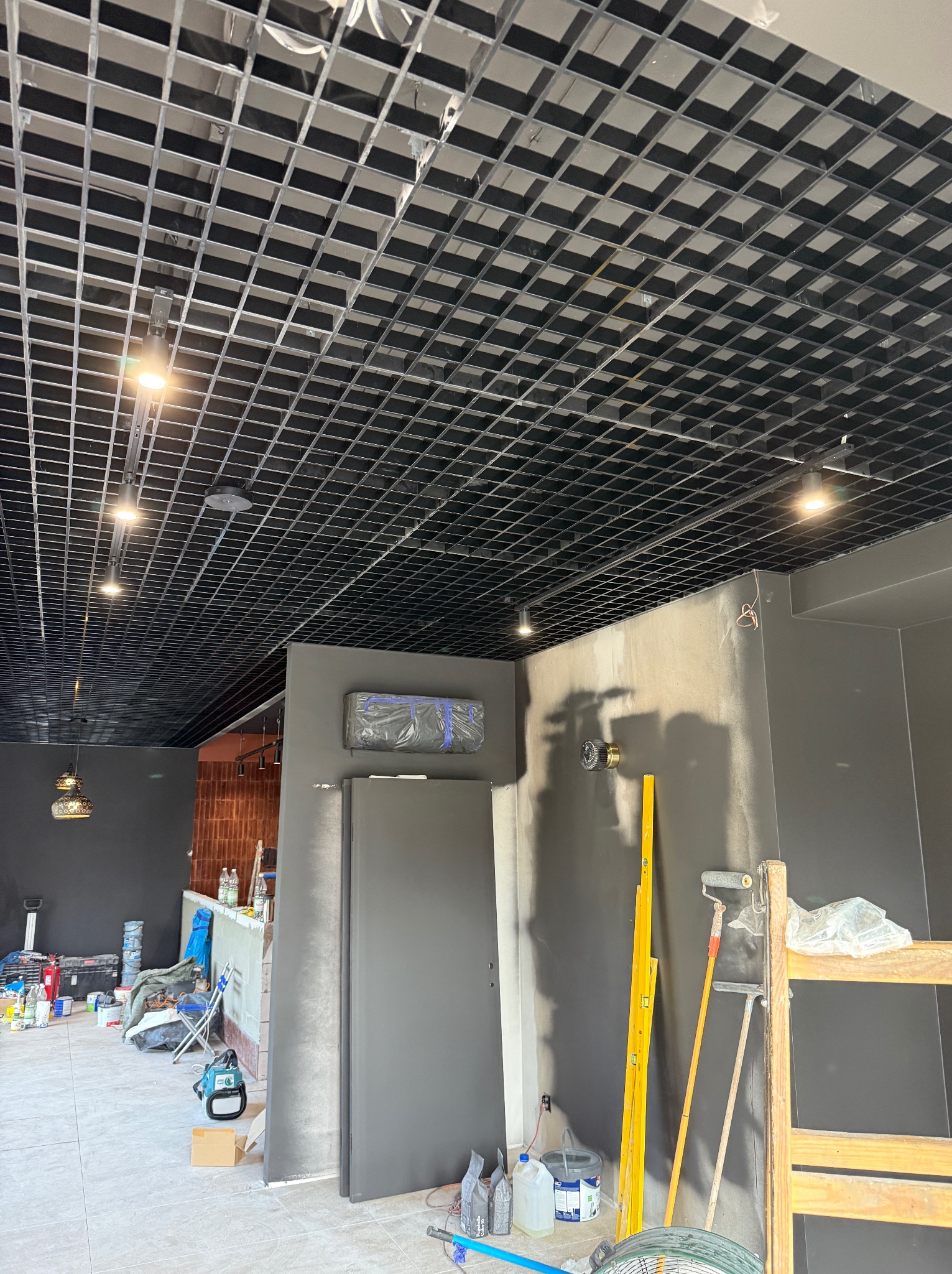 Sufit rastrowy z oświetleniem punktowym w lokalu komercyjnym w trakcie remontu. Widoczne drabina, poziomica i narzędzia malarskie.