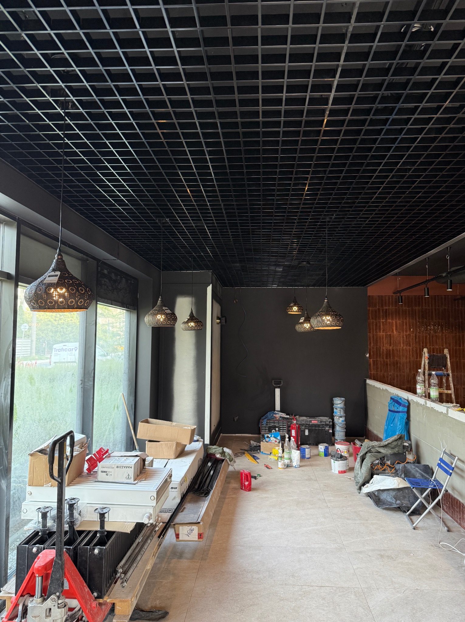 Wnętrze w trakcie aranżacji: wiszące lampy z ażurowymi kloszami, czarny sufit rastrowy, narzędzia i materiały budowlane. Widok na okno i ulicę.