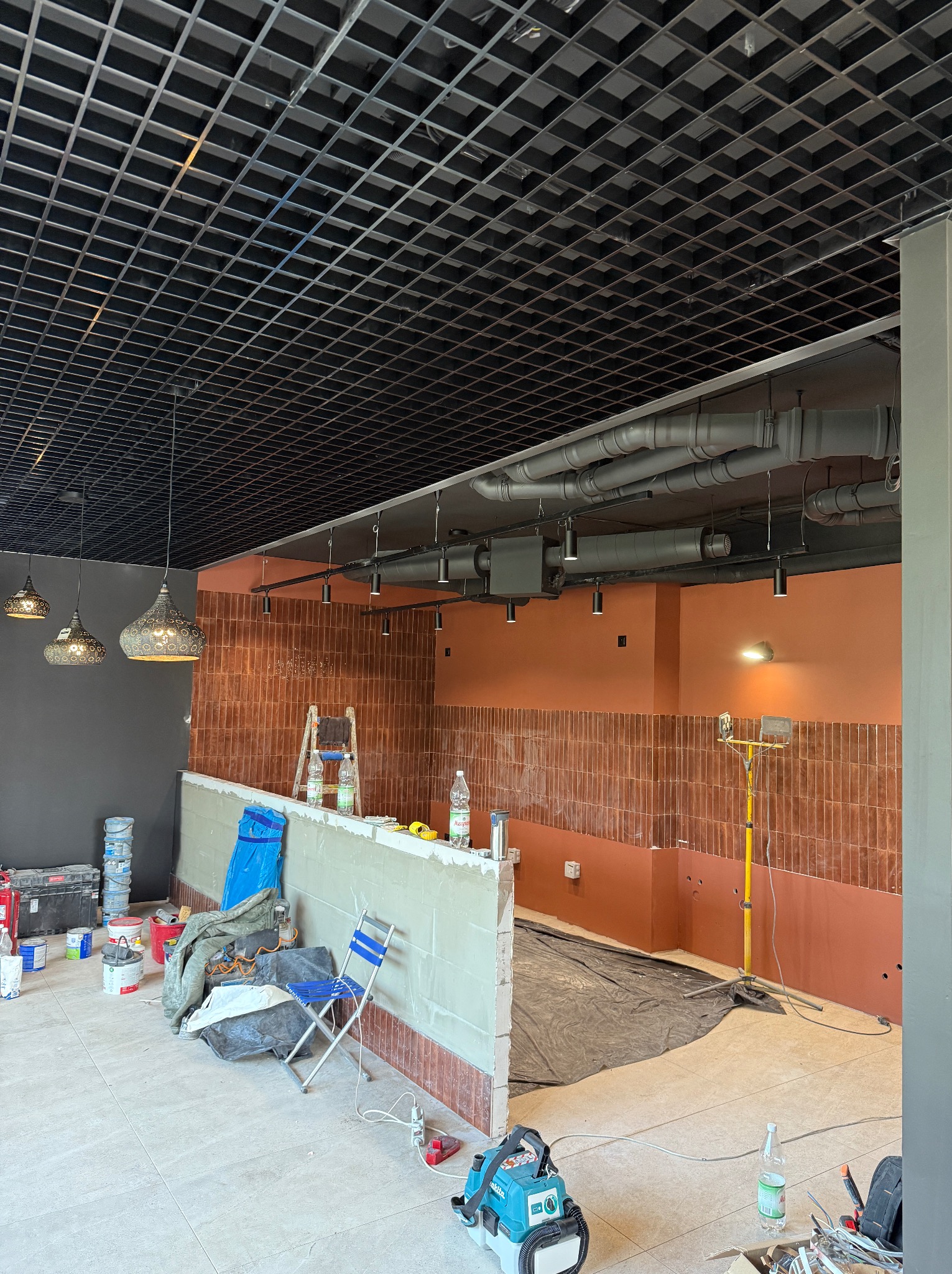 Remont lokalu: sufit rastrowy, rury wentylacyjne, lampy wiszące i punktowe. Ściany w terakocie, częściowo wykończone, narzędzia pracy, drabina.