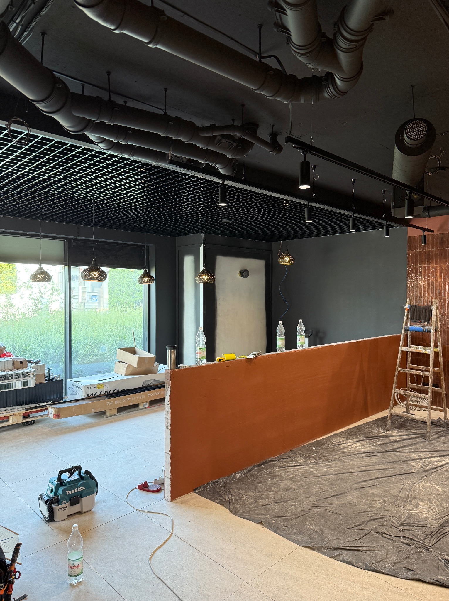 Wnętrze biura w trakcie remontu: widoczne rury na suficie, nowe oświetlenie, pomalowane ściany na szaro, zabezpieczona podłoga folią i drabina oparta o ścianę.