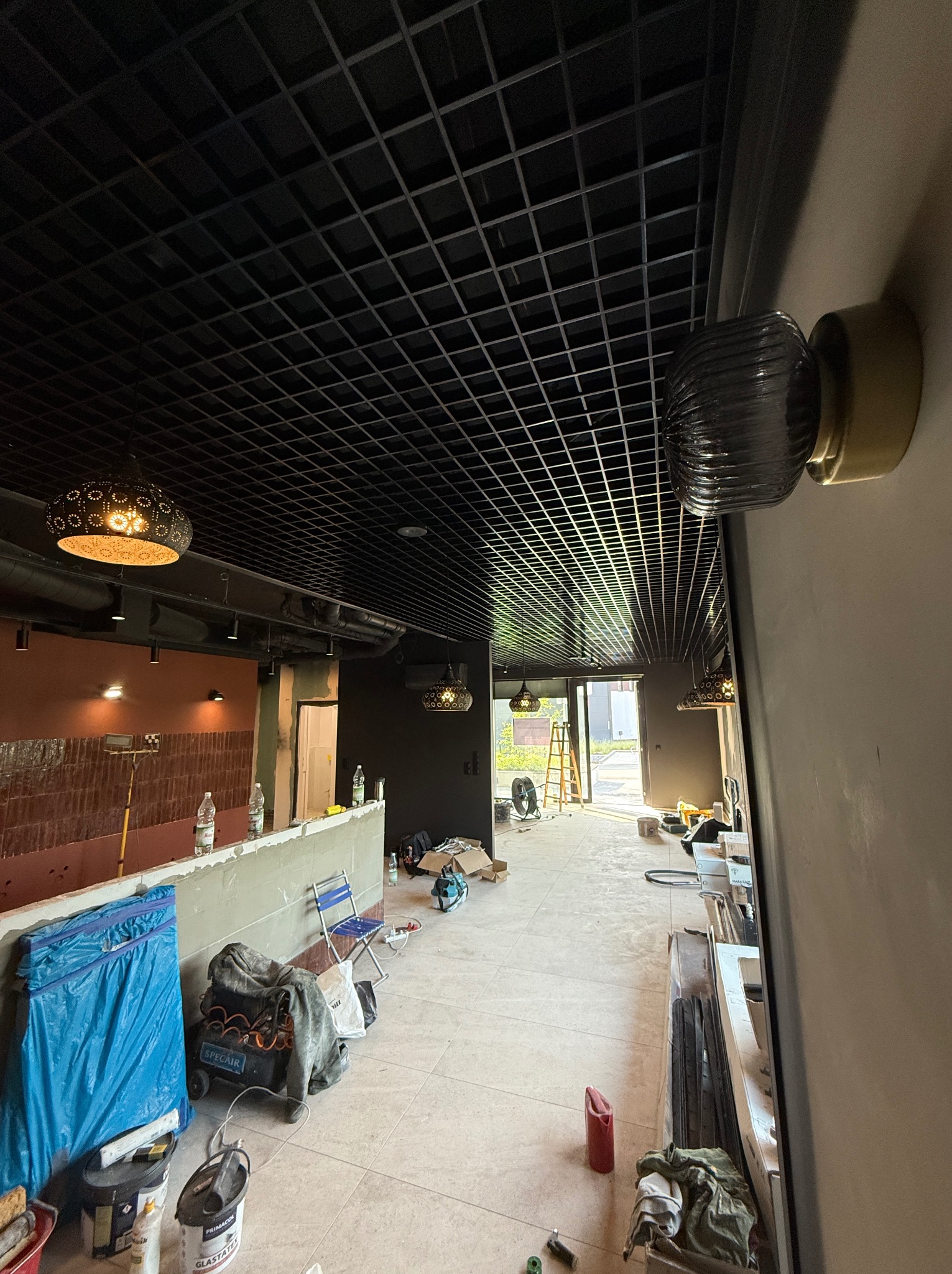 Montaż sufitu rastrowego z oświetleniem w lokalu. Widoczna instalacja elektryczna, lampy wiszące i naścienne. Prace wykończeniowe w trakcie realizacji.