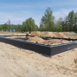 Fundament domu w trakcie budowy, z widoczną izolacją przeciwwilgociową i nasypanym piaskiem wewnątrz, drzewa w tle.