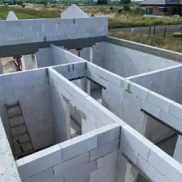 Wnętrze w surowym stanie domu z białych bloczków, widok z góry na wznoszone ściany działowe i nośne, z drewnianą drabiną opartą o ścianę.