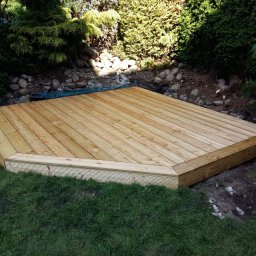 ADAM WOJCIECHOWSKI BRAMYPRO - Drewniany taras w ogrodzie, wykonany z jasnych desek, otoczony zielenią i kamieniami. Naturalne światło podkreśla strukturę drewna i harmonię z otoczeniem.