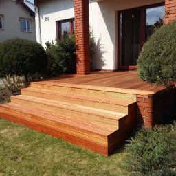 ADAM WOJCIECHOWSKI BRAMYPRO - Nowoczesny taras drewniany z trzema schodkami, wykończony cegłą klinkierową, z widokiem na dom z numerem 36 i zadbany ogród. Słoneczny dzień.