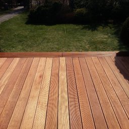 ADAM WOJCIECHOWSKI BRAMYPRO - Nowy taras z desek w kolorze naturalnego drewna, z widokiem na trawnik i ścieżkę w ogrodzie. Deski ułożone równolegle, widoczne delikatne słoje drewna i mocowania.