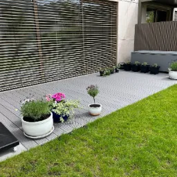 Nowoczesny taras kompozytowy w szarym odcieniu, z donicami z kwiatami i skrzynią ogrodową, przylegający do budynku z żaluzjami zewnętrznymi.