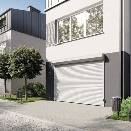 ADAM WOJCIECHOWSKI BRAMYPRO - Nowoczesna brama garażowa rolowana, biała, zamontowana w budynku o minimalistycznej architekturze. Widoczne drzewa i elementy zagospodarowania terenu.