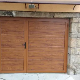 Dwuskrzydłowa brama garażowa w odcieniach brązu, z poziomymi panelami imitującymi drewno, wkomponowana w kamienne obramowanie wjazdowe.