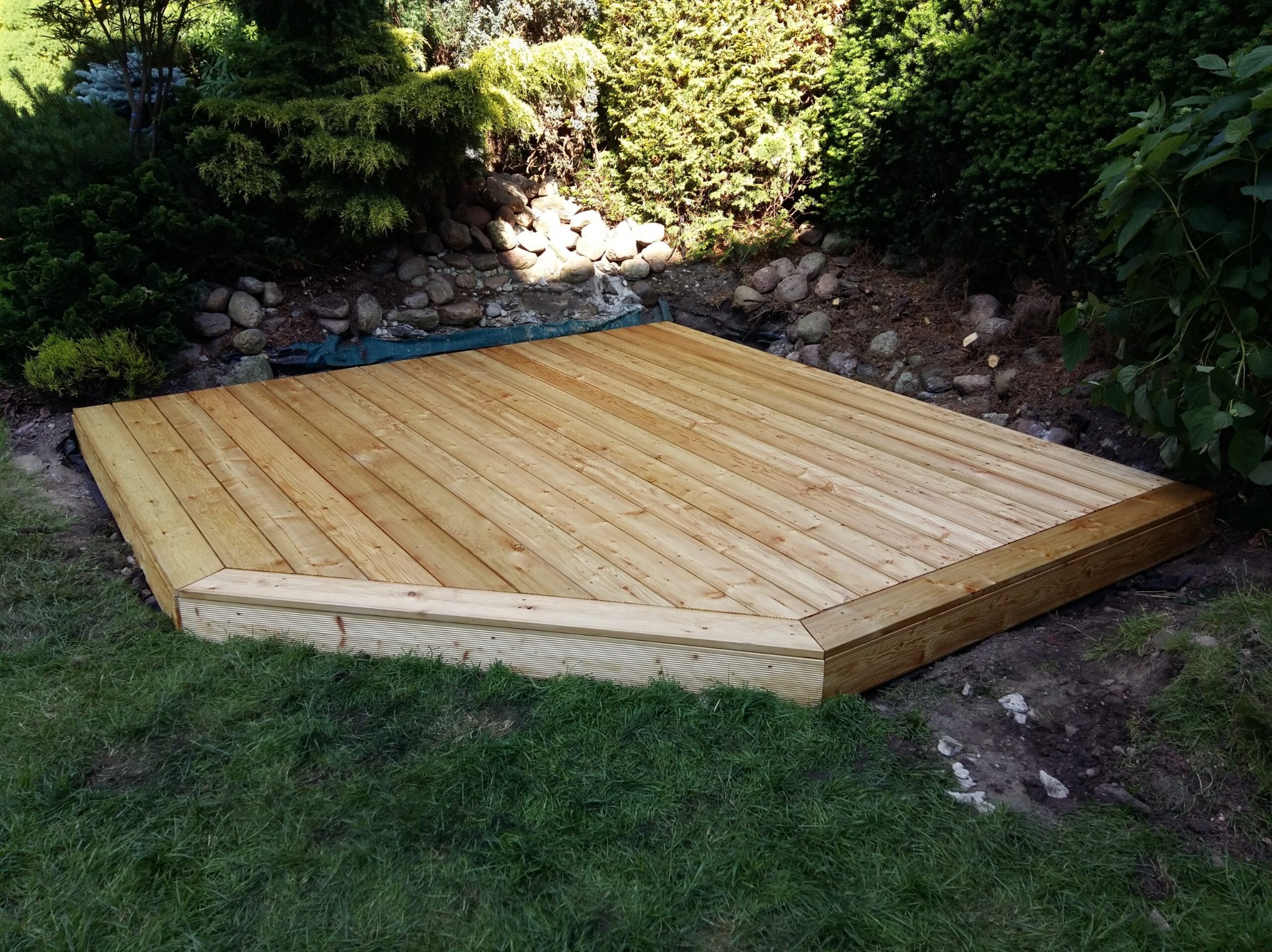 Drewniany taras w ogrodzie, wykonany z jasnych desek, otoczony zielenią i kamieniami. Naturalne światło podkreśla strukturę drewna i harmonię z otoczeniem.