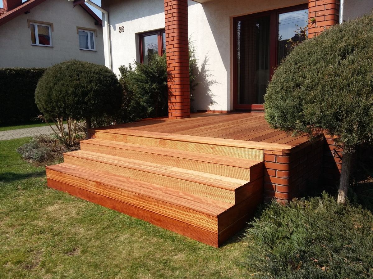 Nowoczesny taras drewniany z trzema schodkami, wykończony cegłą klinkierową, z widokiem na dom z numerem 36 i zadbany ogród. Słoneczny dzień.