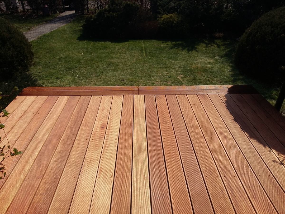 Nowy taras z desek w kolorze naturalnego drewna, z widokiem na trawnik i ścieżkę w ogrodzie. Deski ułożone równolegle, widoczne delikatne słoje drewna i mocowania.