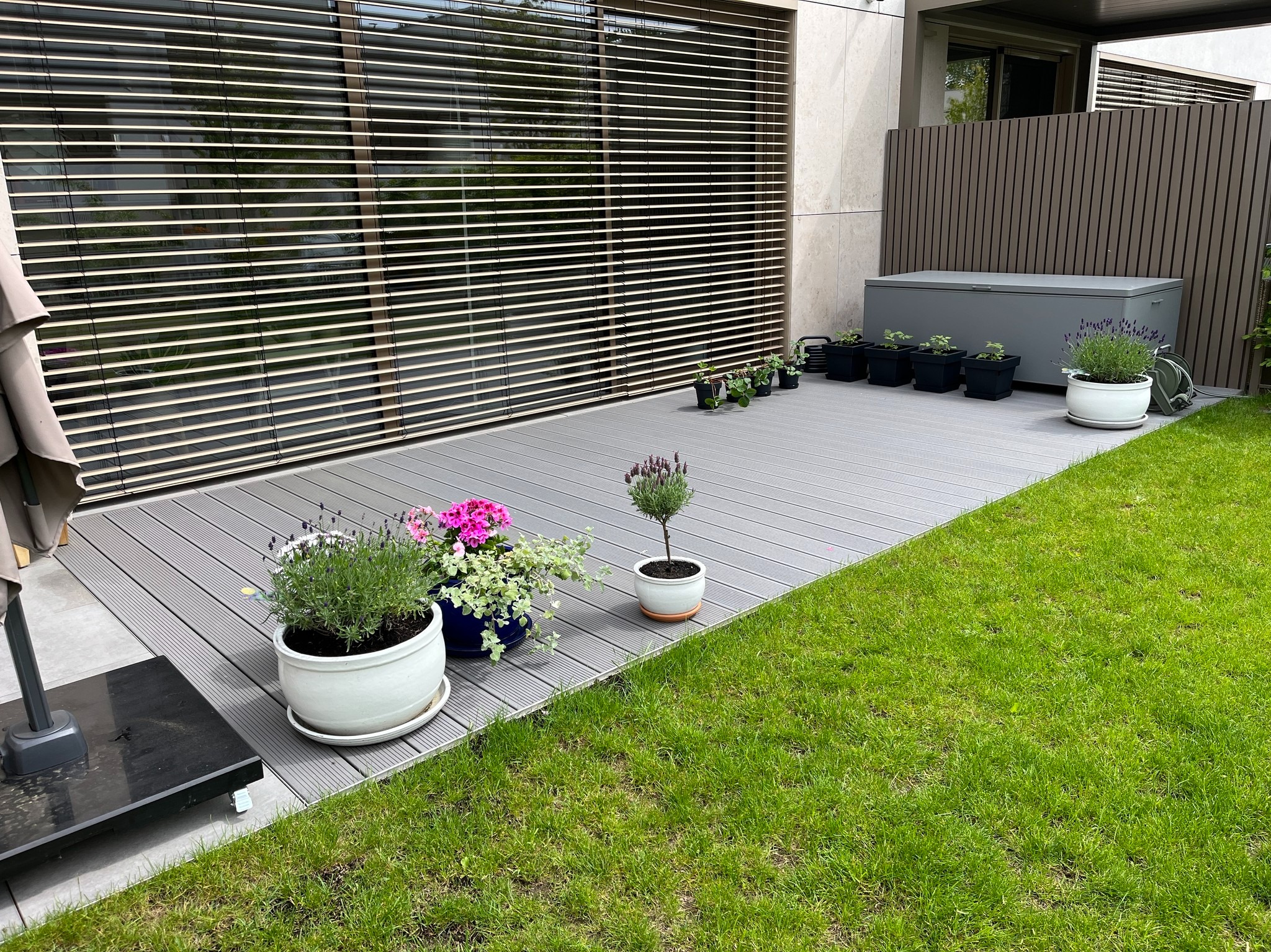 Nowoczesny taras kompozytowy w szarym odcieniu, z donicami z kwiatami i skrzynią ogrodową, przylegający do budynku z żaluzjami zewnętrznymi.
