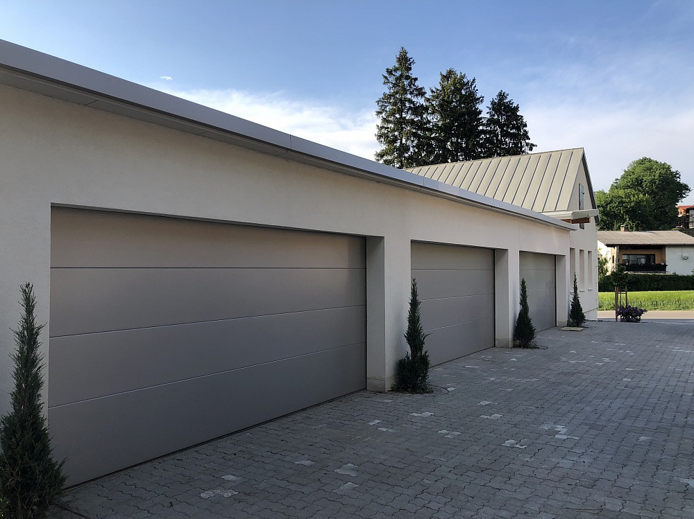 Nowoczesny budynek garażowy z trzema szarymi bramami segmentowymi, ozdobiony smukłymi tujami. Kostka brukowa na podjeździe i budynek mieszkalny w tle.
