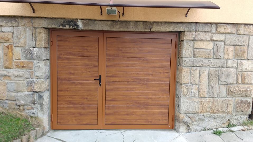 Dwuskrzydłowa brama garażowa w odcieniach brązu, z poziomymi panelami imitującymi drewno, wkomponowana w kamienne obramowanie wjazdowe.