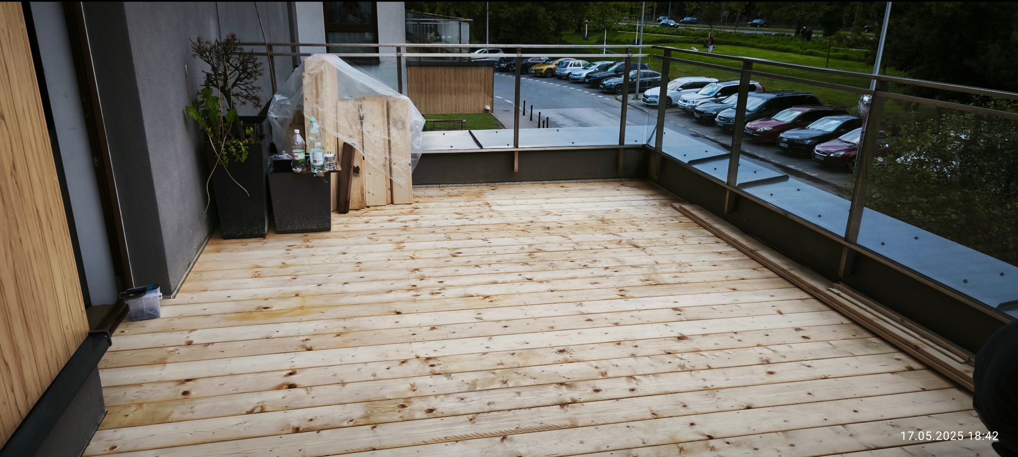 Jasny, drewniany taras z metalową balustradą i szklanymi panelami na tle parkingu. Roślina w donicy i zapakowane deski w rogu. Widok na zewnątrz.