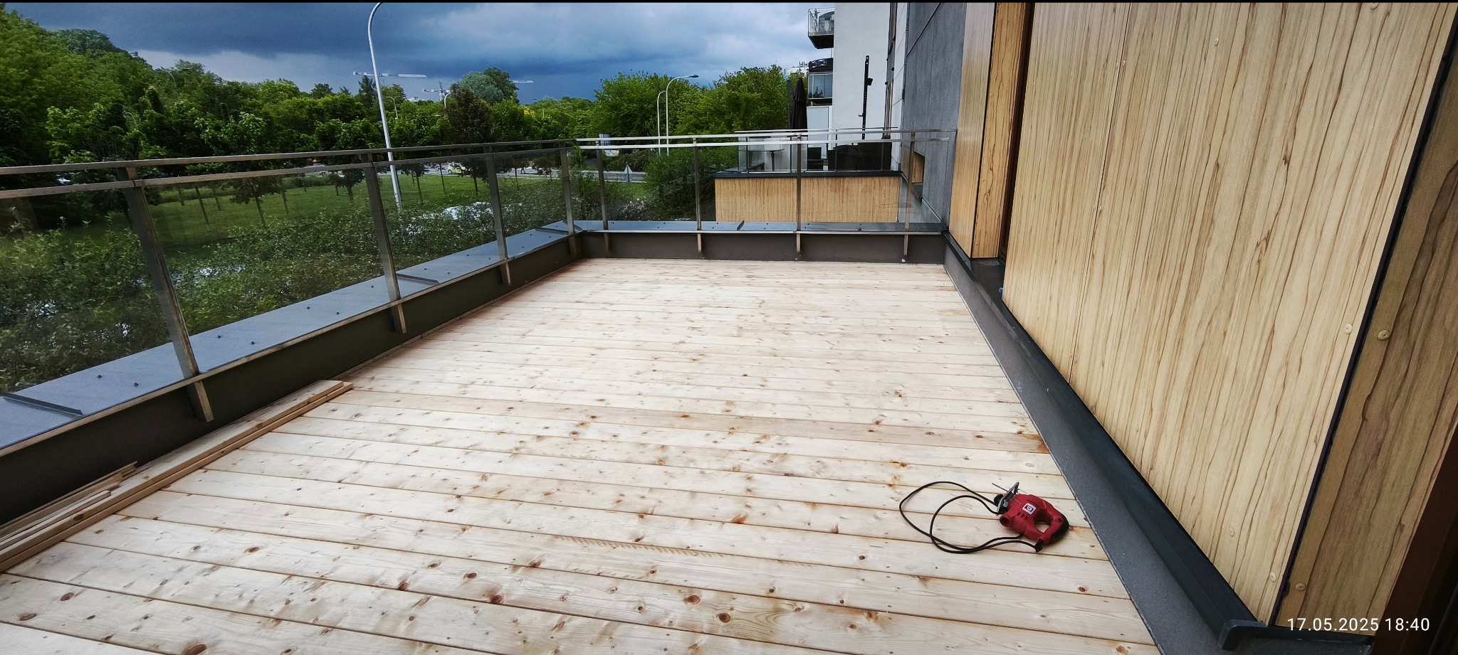 Jasny taras z desek z narzędziem i szklaną balustradą, widok na zieleń. Deski ułożone równolegle, na części tarasu leżą deski do montażu. W tle pochmurne niebo.